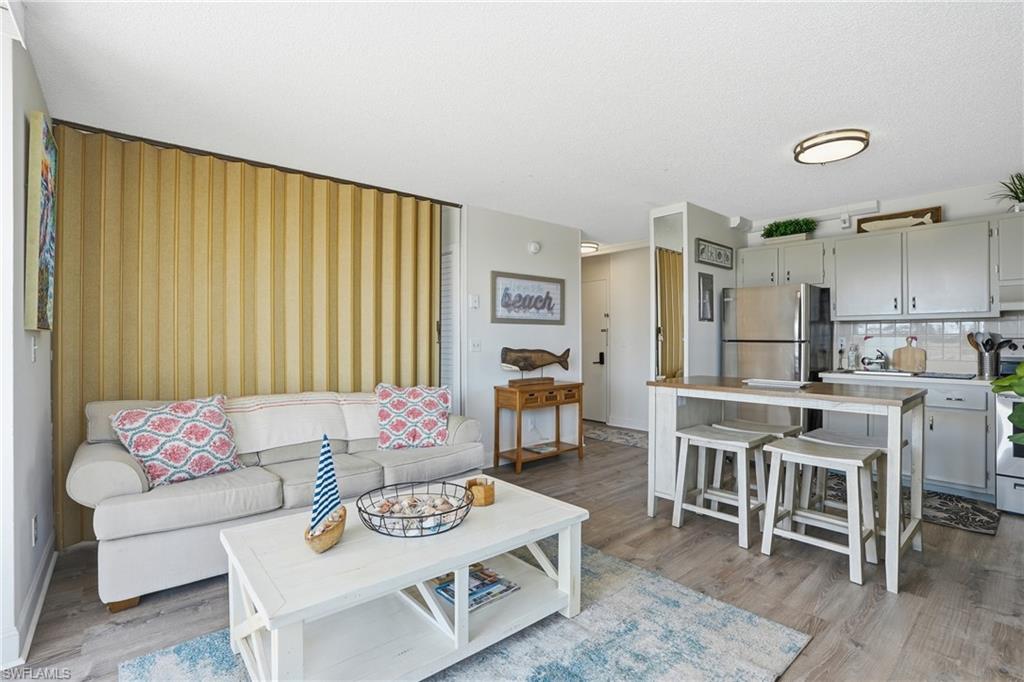 Open-concept living space featuring wood-finish flooring, a kitchen island with seating, stainless steel appliances, and light gray cabinetry - Virtually Edited Image