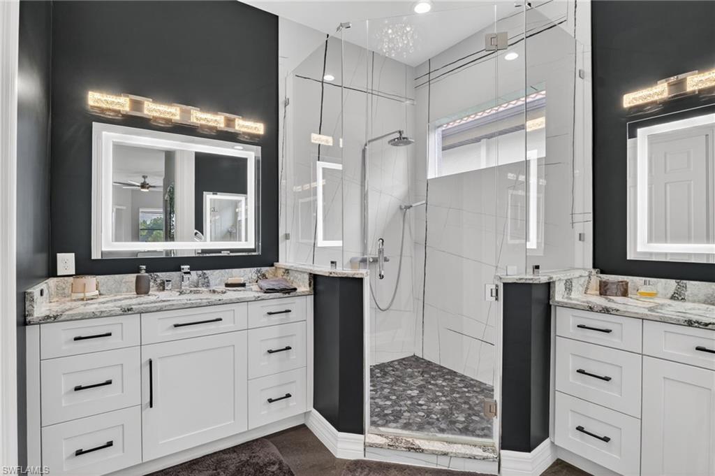 Spacious bathroom featuring a glass-enclosed shower with pebble-finish flooring, dual vanities with white cabinetry and stone countertops, and illuminated LED heated, anti-fog, dimmable, tone adjustable mirrors