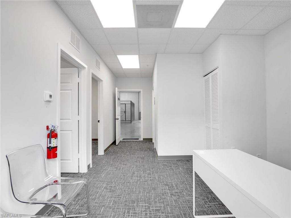 Hallway featuring patterned grey carpet, white walls, multiple doorways, a white built-in desk, and drop ceiling with integrated lighting