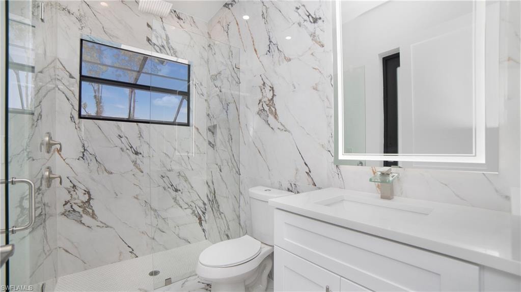 Bathroom featuring full-height marble-style tiled walls, a glass-enclosed shower with a rainfall showerhead, a single vanity with a white countertop and base cabinetry, and an illuminated mirror