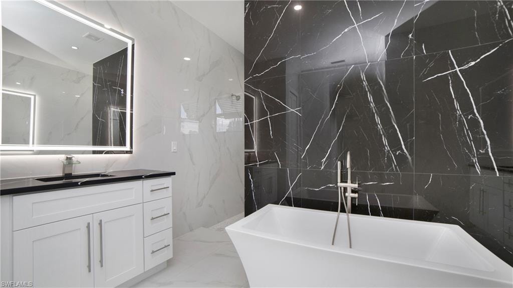 Modern bathroom featuring a standalone soaking tub with floor-mounted faucet, black and white veined tile walls, a white vanity with shaker-style cabinetry, a dark countertop, and an illuminated mirror