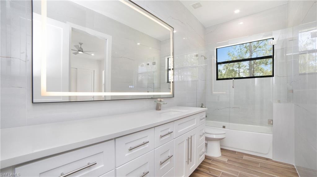 Contemporary bathroom featuring a spacious vanity with white cabinetry and a light-toned countertop, an illuminated rectangular mirror, a white bathtub with a glass shower panel, and wood-finish tile flooring