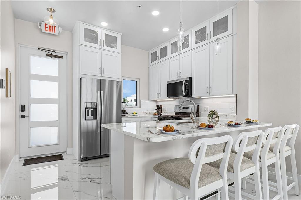 Modern kitchen featuring white shaker cabinetry with glass-front uppers, stainless steel appliances, a white subway tile backsplash, and a white countertop island with seating - Virtually Edited Image