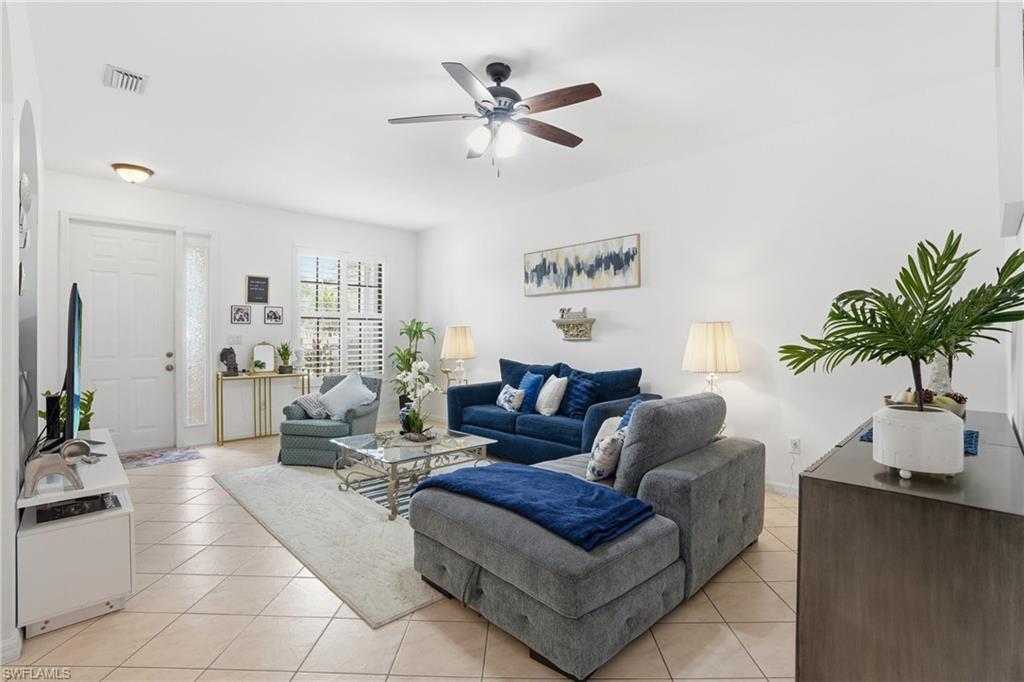 Bright living area featuring neutral tile flooring, white walls, a ceiling fan with integrated lighting, and a white entry door with a sidelight