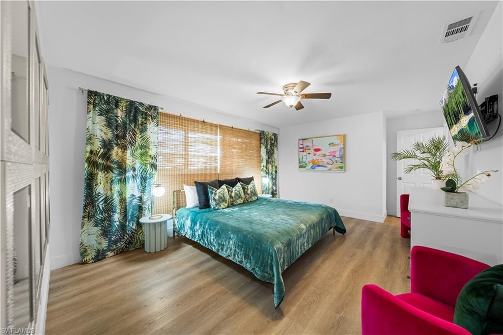 Bright bedroom featuring light wood-finish flooring, a ceiling fan, and decorative window treatments including bamboo blinds and patterned drapes