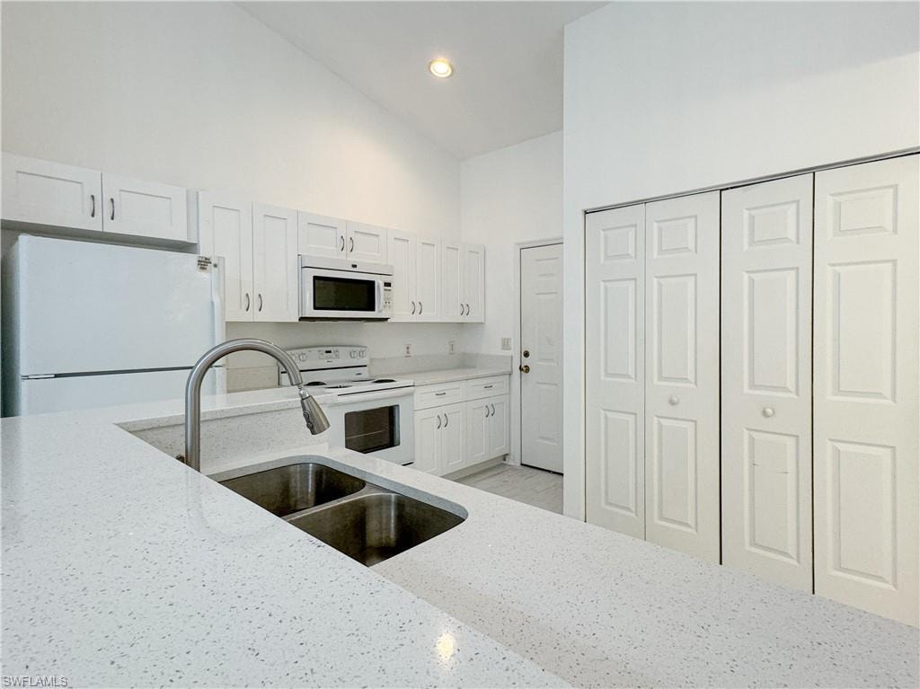 Kitchen with white cabinetry, sink, light stone counters, high vaulted ceiling, and white appliances