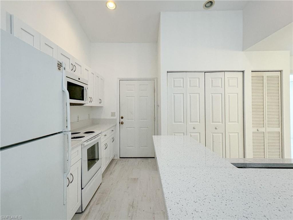 Kitchen featuring light stone counters, white appliances, light hardwood / wood-style floors, a towering ceiling, and white cabinetry