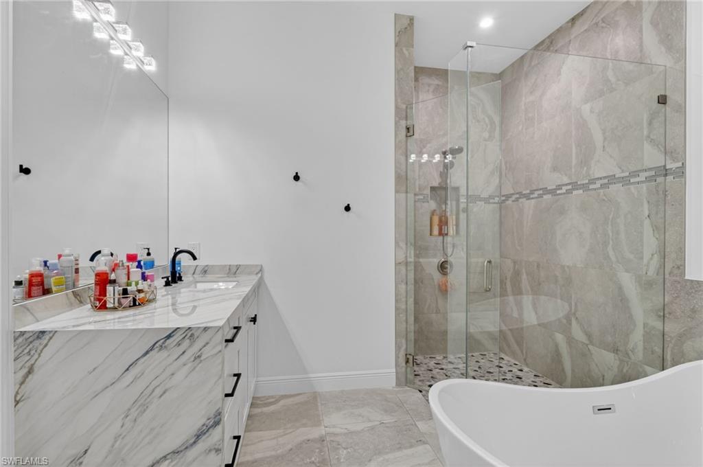 Bathroom featuring a frameless glass shower enclosure with floor-to-ceiling tile, a freestanding soaking tub, a single vanity with a stone countertop, a large mirror, and recessed lighting