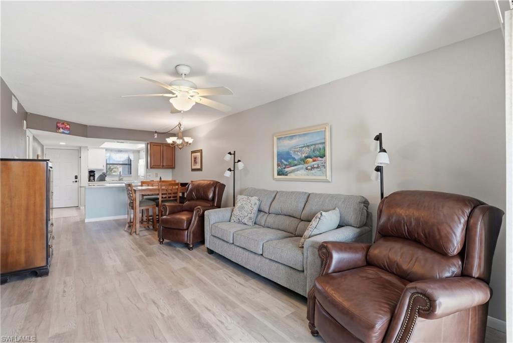 Open concept living area featuring wood-finish flooring, a ceiling fan with integrated lighting, and a partial wall separating a kitchen with light wood cabinetry and a breakfast bar - Virtually Edited Image