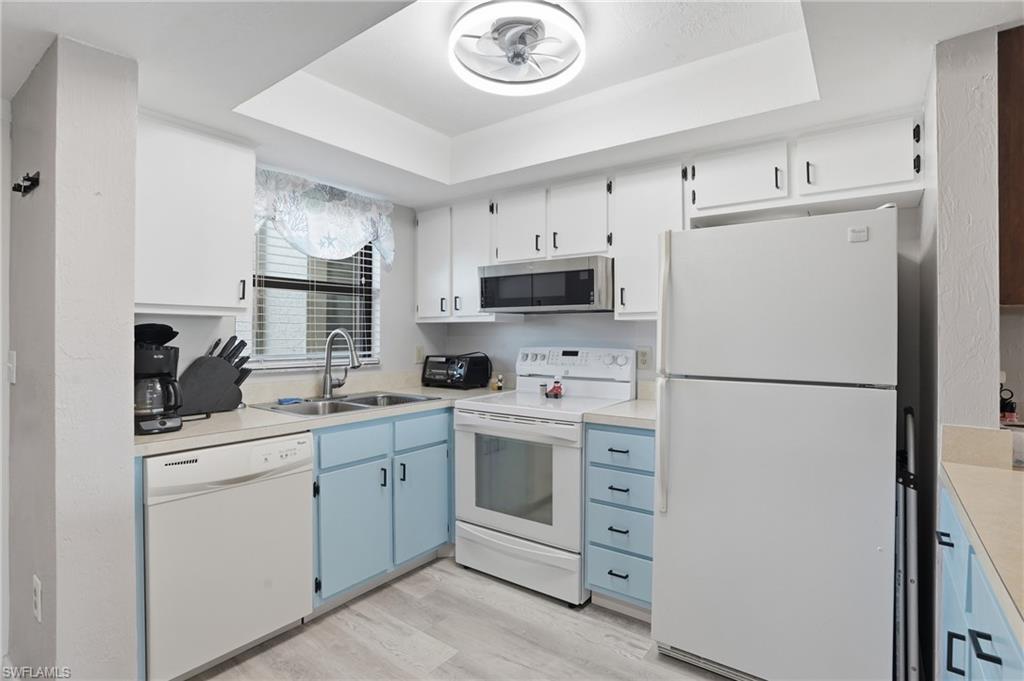 Galley kitchen featuring white appliances, a double basin stainless steel sink, wood-finish flooring, white upper cabinetry, and light blue lower cabinetry - Virtually Edited Image