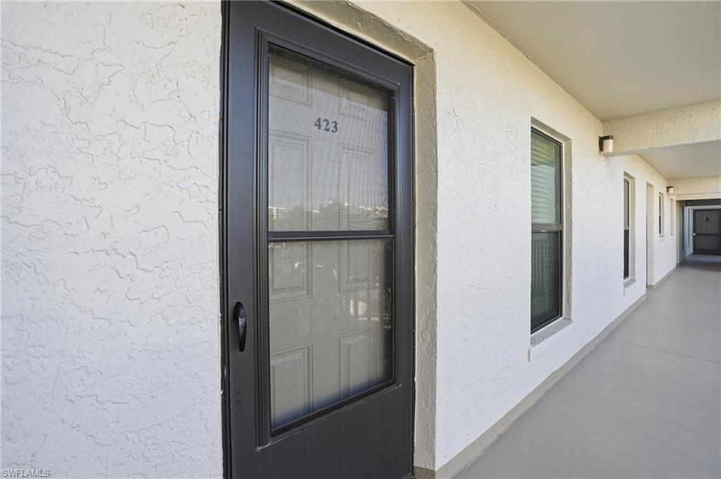 Unit entrance featuring a dark-framed screen door with house number 423, a textured white stucco exterior, and a covered outdoor corridor with multiple windows and exterior lighting fixtures - Virtually Edited Image