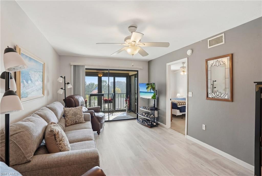 Spacious living area featuring light wood-finish flooring, a ceiling fan, and access to an outdoor balcony through sliding glass doors - Virtually Edited Image