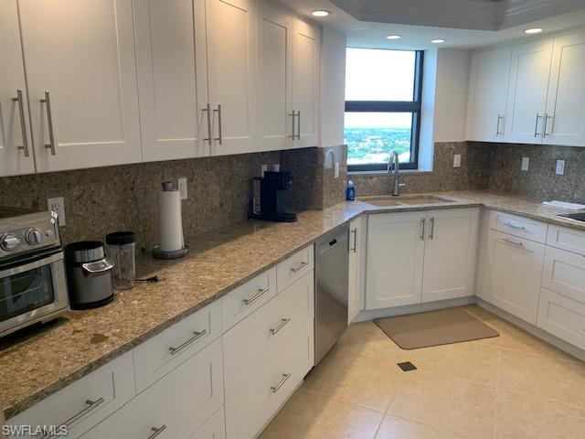 Kitchen featuring dishwasher, sink, light stone countertops, decorative backsplash, and white cabinets