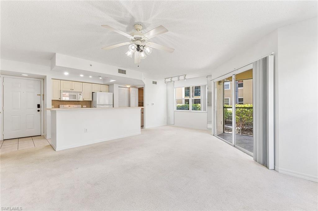 Spacious living area featuring light-toned carpeting, a ceiling fan with integrated lighting, and white baseboards, exterior slider to the lanai, and tile floors in the kitchen area.