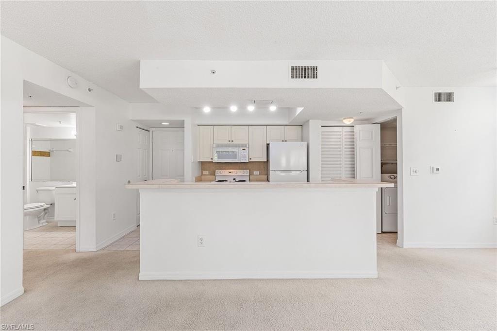 Kitchen with an eat-at counter, light cabinetry, white appliances, and a tiled backsplash, and tile floors in the kitchen and carpet in the living areas.