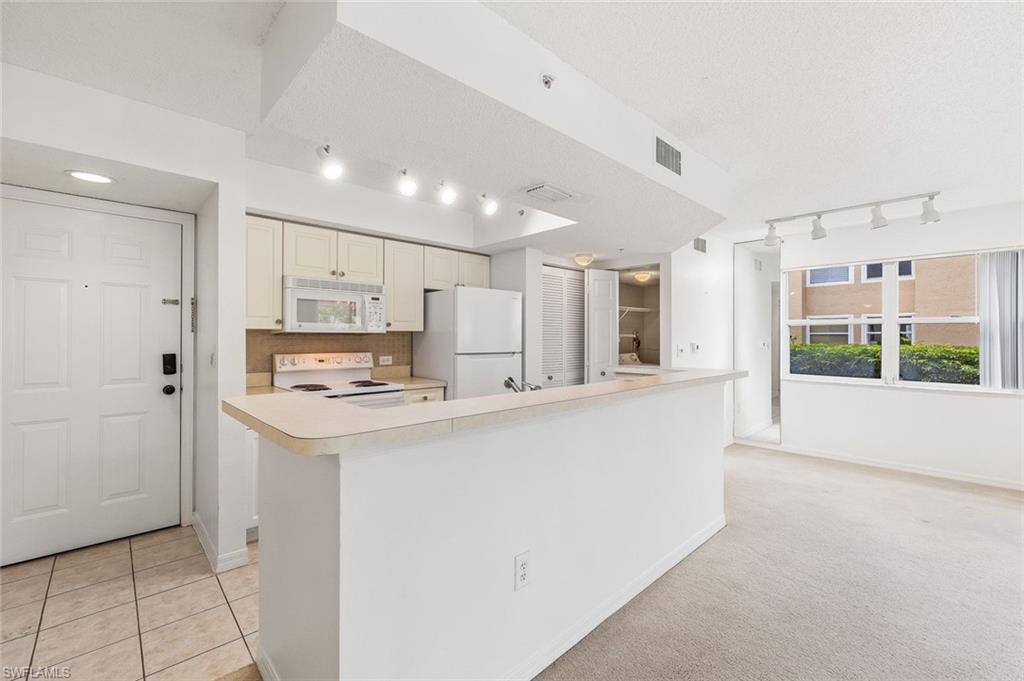 Open-concept living area featuring a kitchen with light-toned cabinetry, white appliances, and track lighting, and tile floors in the kitchen and carpet in the living areas.
