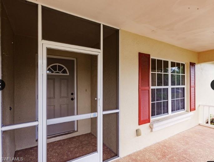 Screened-in porch entry featuring a decorative front door with an arched transom, a large window with red louvered shutters, and a textured concrete floor