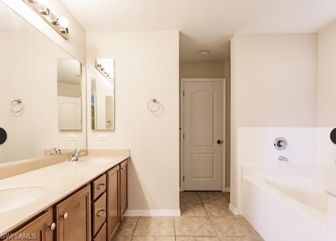 Bathroom featuring a double vanity with light wood cabinetry, a built-in bathtub with white tile surround, and neutral floor tiling