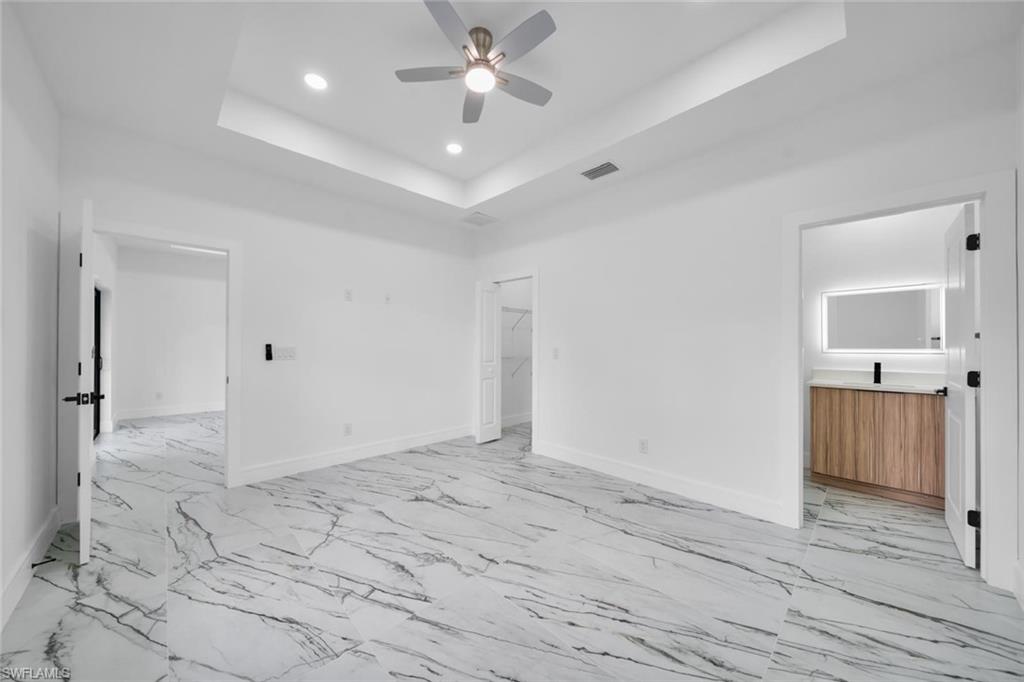 Spacious room featuring large format tile flooring, a tray ceiling, recessed lighting, a ceiling fan, and access to an ensuite bathroom with a modern vanity and illuminated mirror