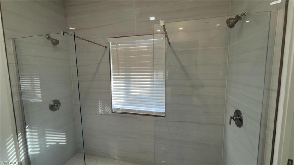 Spacious shower enclosure featuring two independent shower heads, clear glass partitions, light-toned horizontal tile work, a window with blinds, and white hexagonal floor tiling