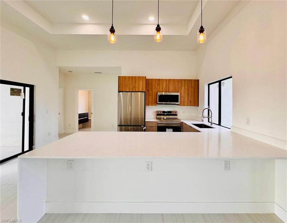 Kitchen featuring a large central island with a light-toned countertop, stainless steel appliances, wood-finish cabinetry, a deep sink with a gooseneck faucet, and recessed lighting with pendant fixtures