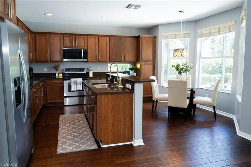 Kitchen featuring wood-finish cabinetry, stainless steel appliances, dark countertops, a central island with an integrated sink, and wood-finish flooring