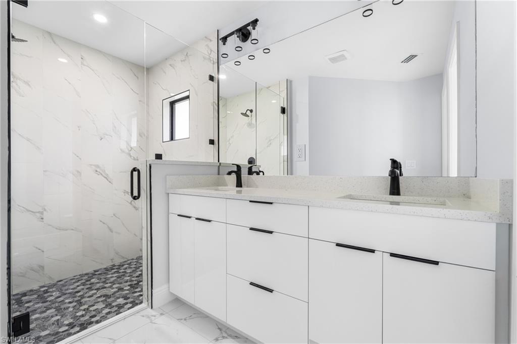 Spacious bathroom featuring a frameless glass shower enclosure with stone mosaic flooring, dual white vanities with integrated sinks, and black hardware throughout