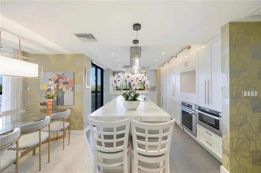 Spacious kitchen featuring a large central island with a light-toned countertop, extensive white cabinetry with gold-toned hardware, double wall ovens, and pendant lighting
