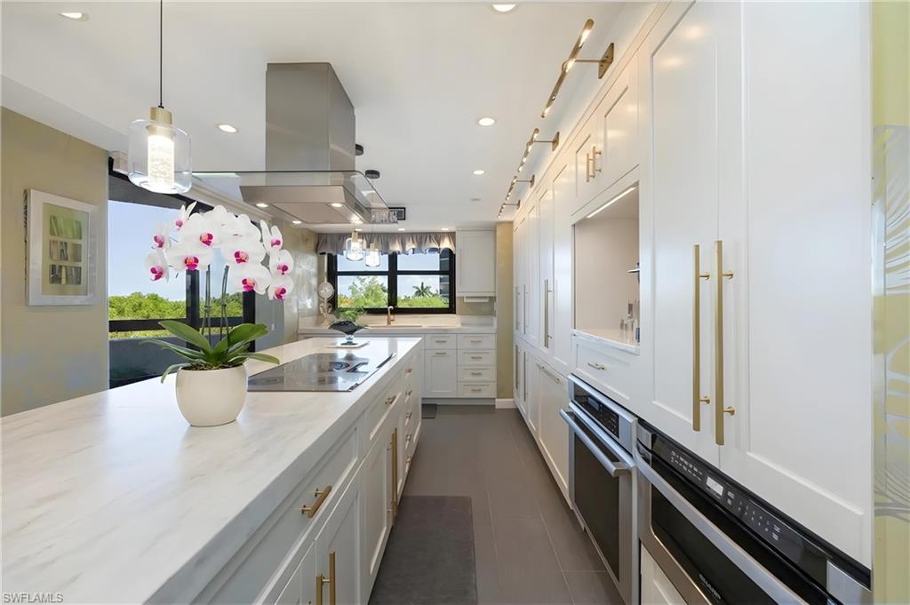 Contemporary kitchen featuring an integrated cooktop on a central island with white stone-finish countertops, extensive white cabinetry with brass hardware, and built-in stainless steel appliances