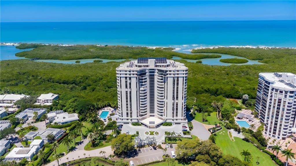High-rise building with a curved façade and multiple balconies, featuring a circular drive, lush green landscaping, and an outdoor swimming pool, all against a backdrop of a clear blue ocean and dense coastal foliage