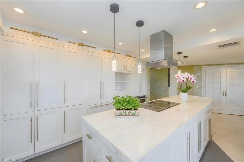 Gourmet kitchen featuring a substantial island with integrated cooktop, stainless steel range hood, white shaker cabinetry with gold-toned hardware, recessed lighting, and pendant lights
