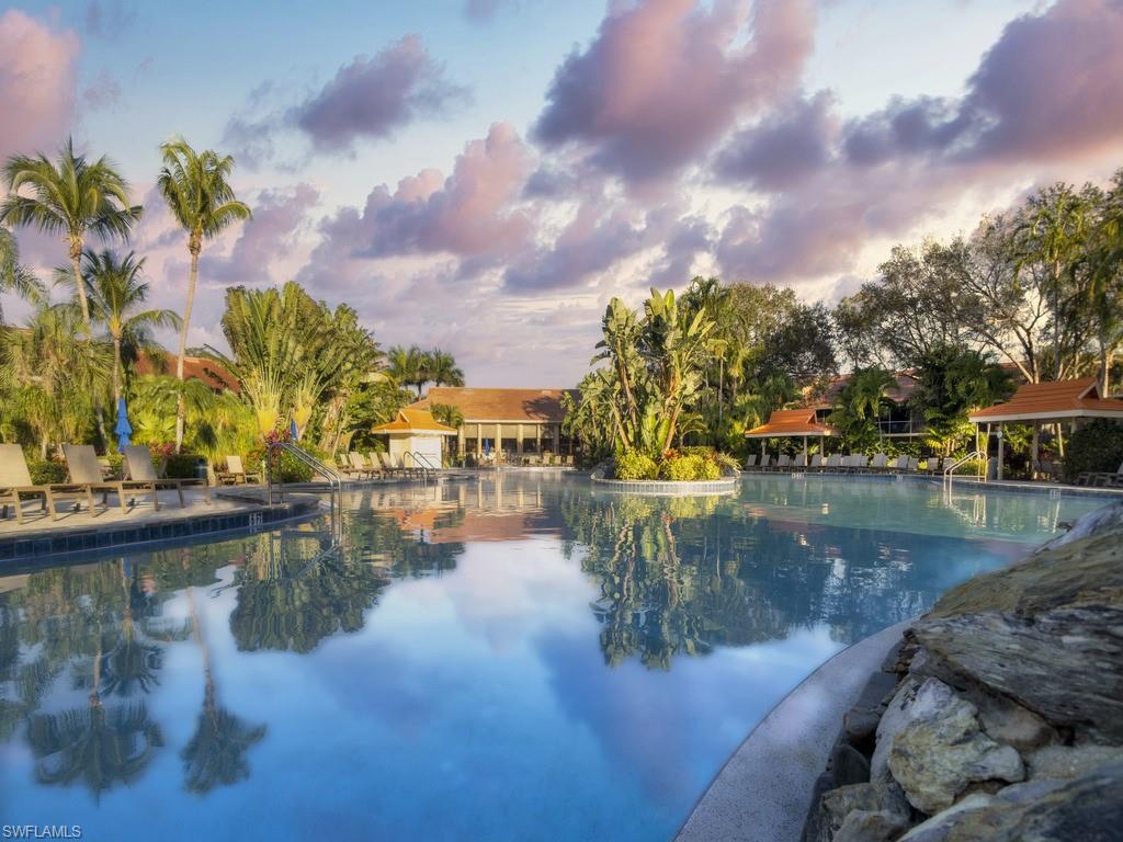 Expansive resort-style swimming pool featuring integrated water features, surrounding lounging areas, mature tropical landscaping, and a distant building with a terra cotta tile roof
