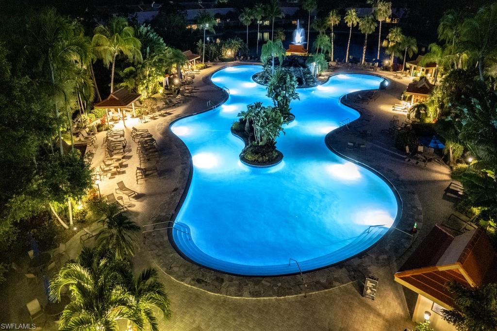 Expansive resort-style pool featuring an irregular shape, illuminated water, multiple integrated islands with lush landscaping, surrounding paved decking, and mature palm trees