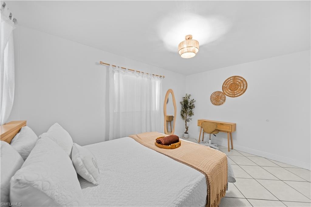 Bedroom featuring light-toned tile flooring, white walls, and a window with sheer curtains on a wooden rod
