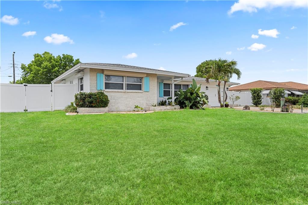 White brick exterior with turquoise shutters, a well-maintained lawn, privacy fencing, and tropical landscaping