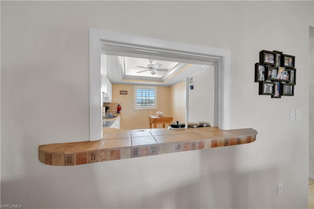 Pass-through window featuring a tiled countertop with a rounded edge, white trim, and a tray ceiling with a ceiling fan in the background
