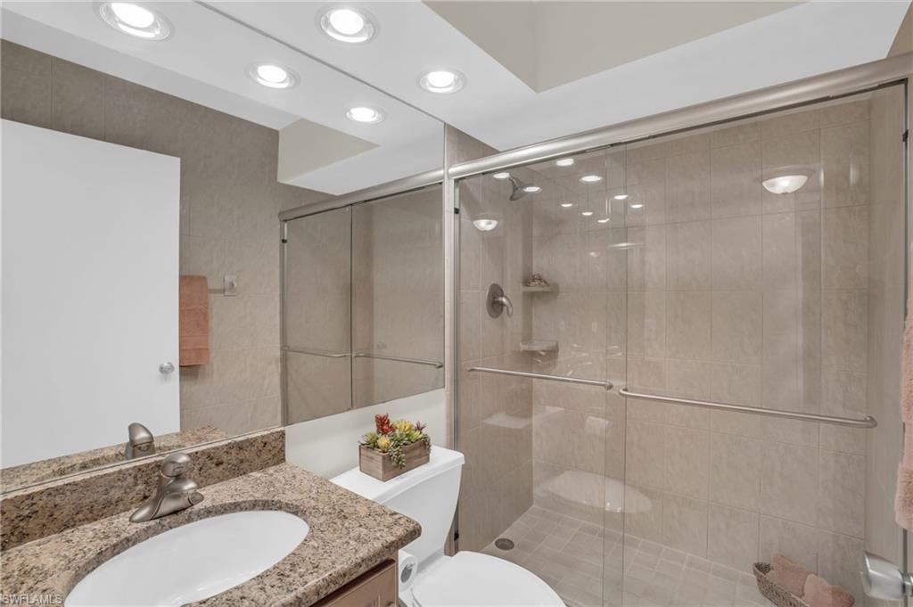 Bathroom featuring a granite-finish vanity with an undermount sink, a full-wall mirror, recessed ceiling lighting, and a glass-enclosed shower with tile surfacing and built-in shelving