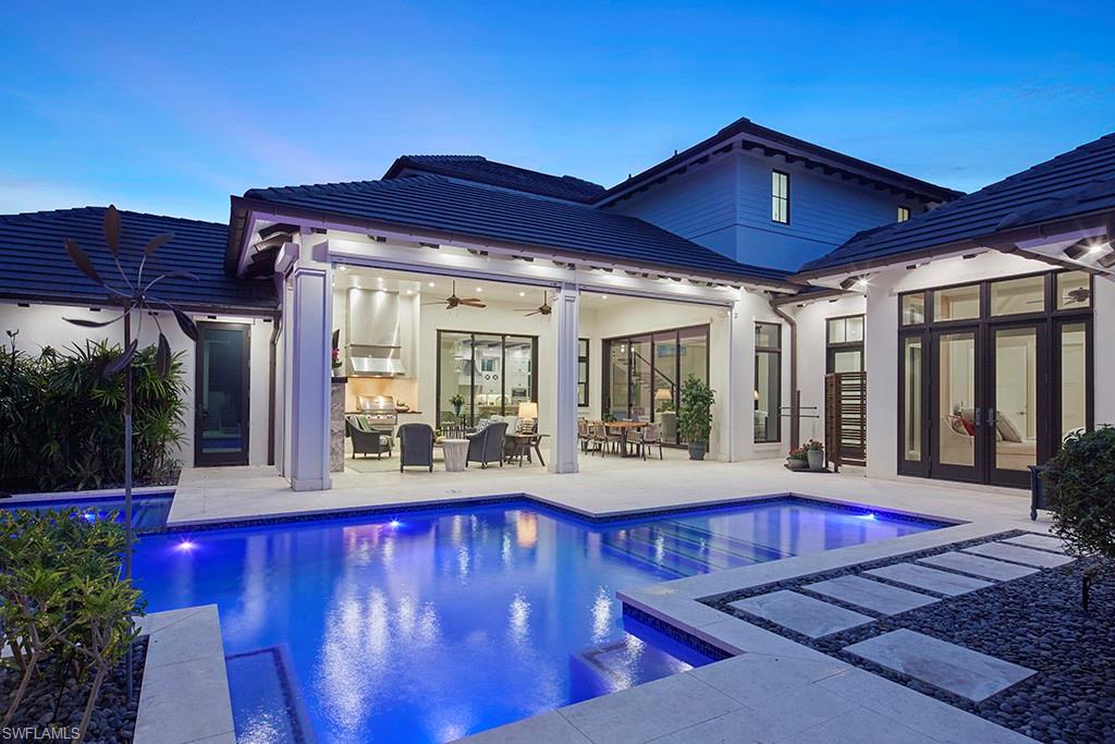 Stunning modern residence showcasing an illuminated resort-style pool with spa, expansive covered loggia with floor-to-ceiling glass doors, multiple sliding glass panels opening to the outdoor entertaining space, sophisticated architectural design with pitched rooflines and dark tile detailing, and pristine travertine or light-colored stone decking surrounding the pool area