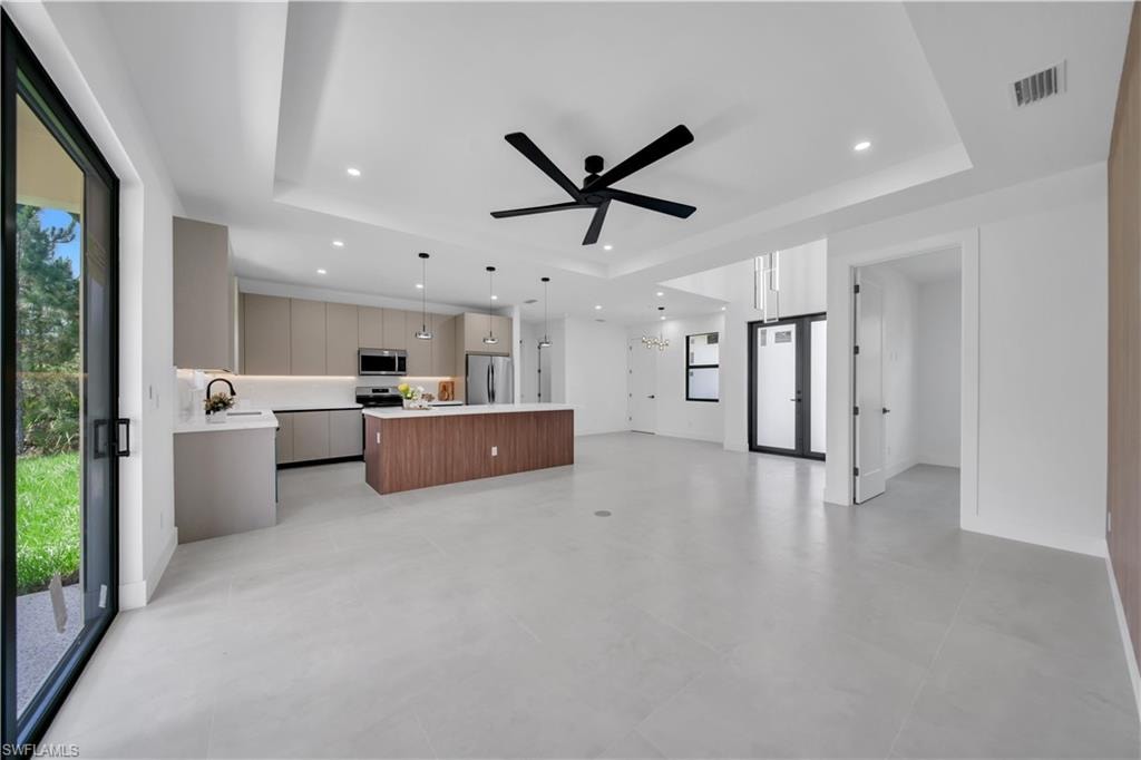 Spacious open-concept living area featuring light-toned tile flooring, a contemporary kitchen with wood-finish island and light grey cabinetry, recessed lighting, a modern ceiling fan, and large sliding glass doors to the exterior
