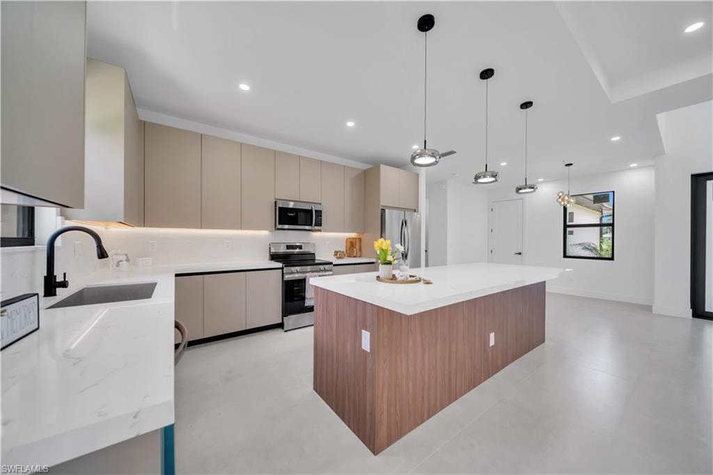 Contemporary kitchen featuring a large central island with a white countertop, matte beige cabinetry, stainless steel appliances, and sleek pendant lighting
