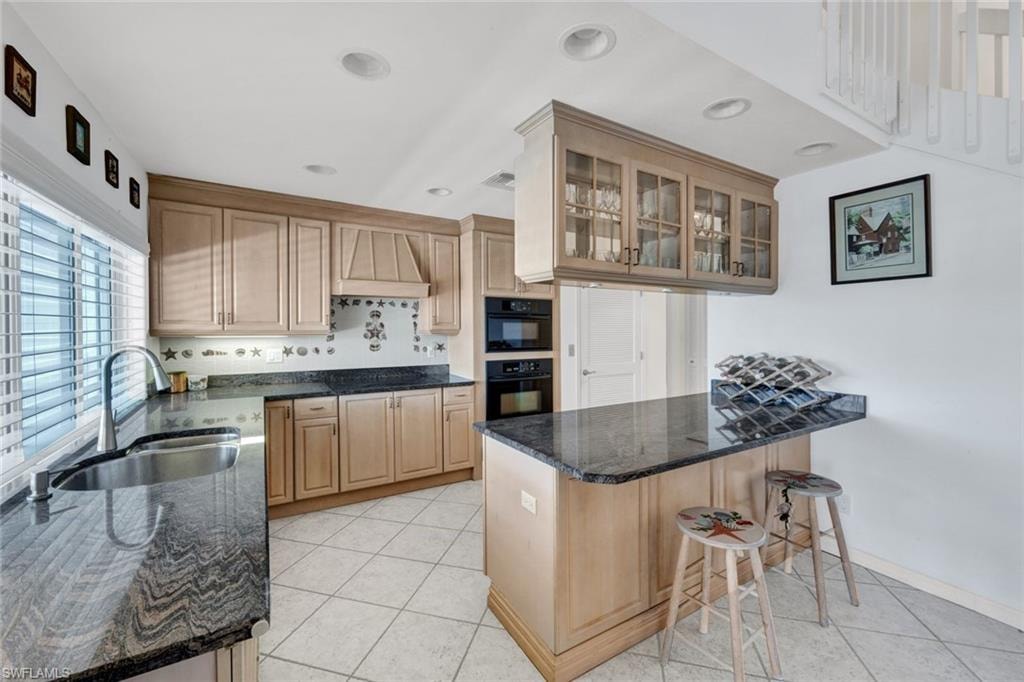 Kitchen featuring light wood cabinetry, dark stone countertops, stainless steel sink, built-in double ovens, and recessed lighting