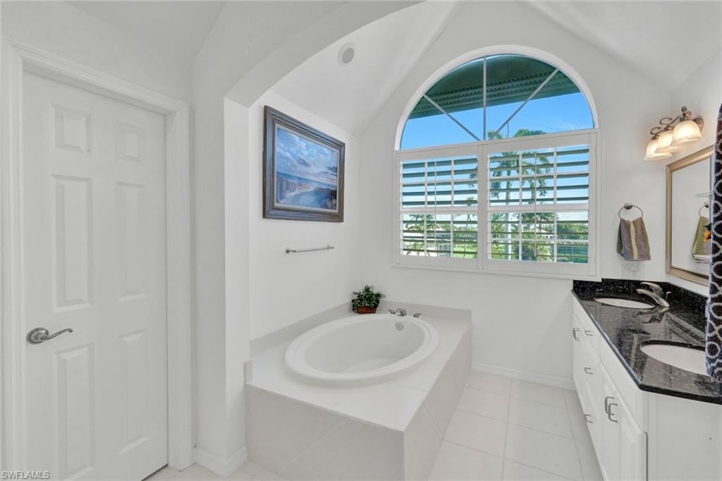 Bathroom featuring an arched window with louvered shutters, a built-in soaking tub, a double vanity with a dark countertop and undermount sinks, and white tile flooring