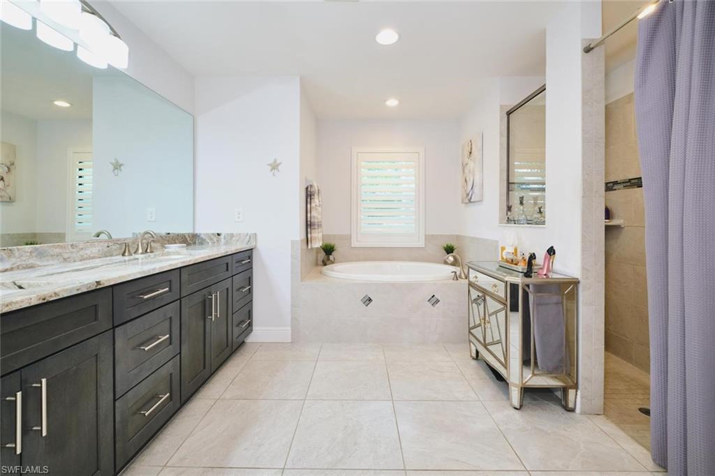 Spacious bathroom featuring a dual vanity with a stone countertop and dark wood-finish cabinetry, a large built-in soaking tub, a separate glass-enclosed shower, and neutral tile flooring