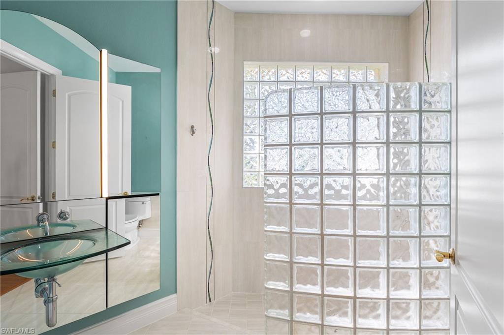 The third full bathroom, a beautifully appointed ensuite, offers a striking sense of refinement with a dramatic glass block feature wall that creates a seamless, walk-in shower experience. Floor-to-ceiling porcelain tile, accented with elegant listello detailing, adds a sophisticated architectural finish, while an expansive glass block window fills the space with radiant natural light. A sleek single glass vanity completes the design, preserving a clean, modern aesthetic with understated luxury.