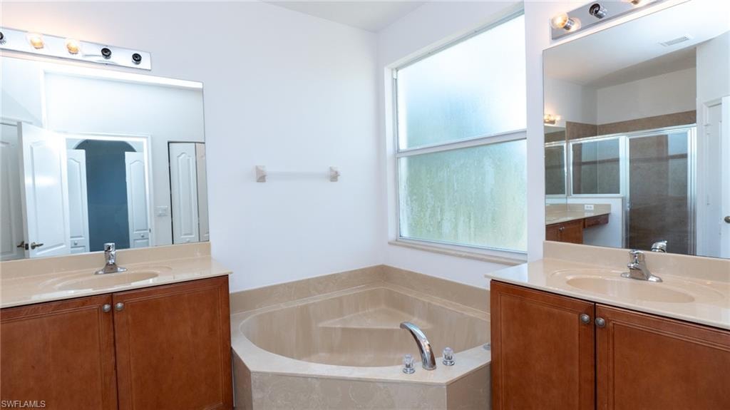 Bathroom featuring a corner soaking tub with deck-mounted fixtures, dual vanity sinks with light-toned countertops, wood-finish cabinetry, and a large frosted window