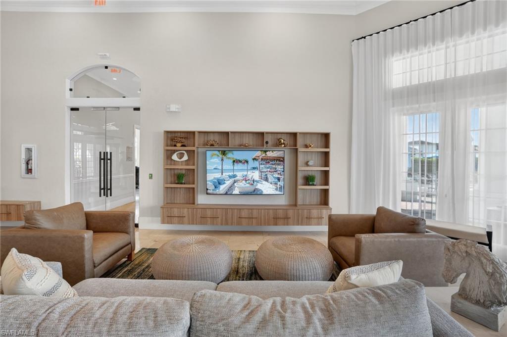 Spacious common area featuring a light wood-finish built-in entertainment center, high ceilings, large windows with sheer white drapery, and an arched entryway with double glass doors