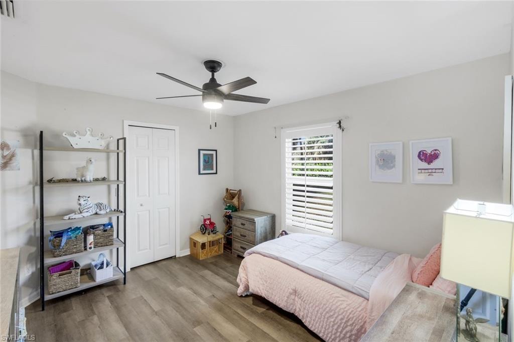 Bedroom featuring wood-finish flooring, a ceiling fan with integrated lighting, a window with plantation shutters, light-toned walls, and a bi-fold closet door