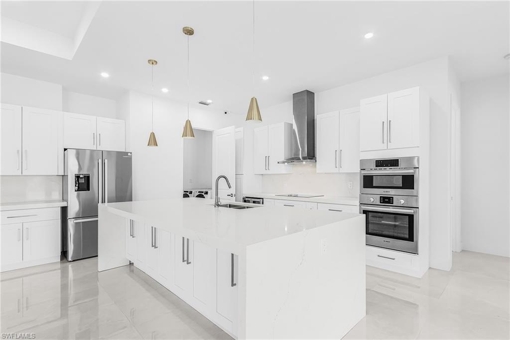 Bright kitchen featuring a large center island with integrated sink, white cabinetry, stainless steel appliances, and sleek pendant lighting