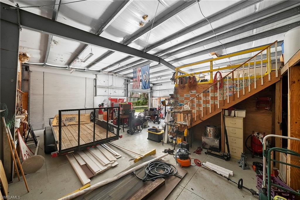 Spacious garage interior featuring industrial-style steel beam construction, two large roll-up doors, concrete flooring, a built-in wooden staircase with a banister, and a loft area with a yellow safety railing
