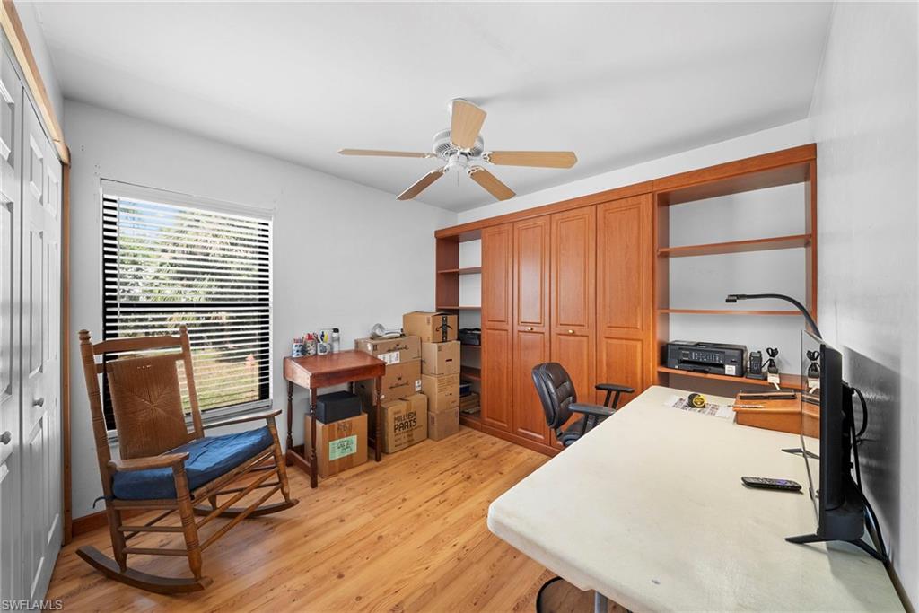 Room featuring wood-finish flooring, a built-in cabinetry unit with enclosed storage and open shelving, a ceiling fan with light fixture, and a window with horizontal blinds